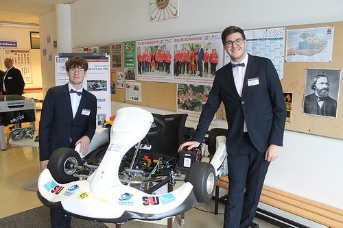Zwei junge Männer in Anzügen stehen neben einem weißen Go-Kart. Das Go-Kart hat die Buchstaben 'SAU' und 'F' darauf. Sie befinden sich in einem Raum mit einer Anschlagtafel und einem Kalender mit dem Jahr 2024.