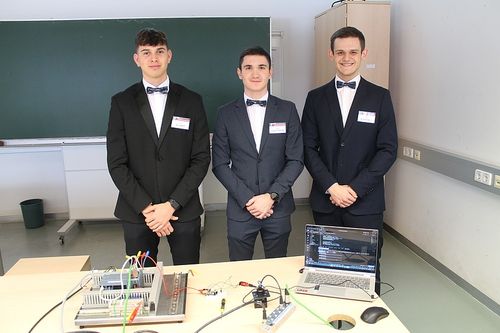 Drei Männer in formellen Anzügen mit Fliegen stehen vor einem Tisch mit Laptop und Kabeln. Hinter ihnen ist ein Klassenzimmer mit einer grünen Tafel und einem Schrank.