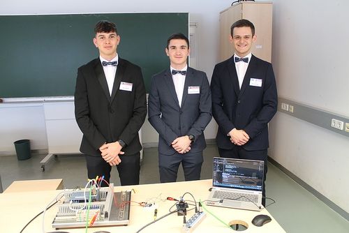 Drei Männer in formellen Anzügen mit Fliegen stehen vor einem Tisch mit Laptop und Kabeln. Hinter ihnen ist ein Klassenzimmer mit einer grünen Tafel und einem Schrank.