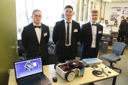 Drei junge Männer in Anzügen und Fliegen stehen in einer Werkstatt. Ein Roboter steht auf einem Tisch, mit einem Laptop, das ein 3D-Modell anzeigt.