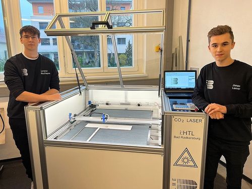 Zwei junge Männer stehen neben einer CO2-Lasermaschine, einer mit Brille, beide lächeln, in einem Raum mit Glasfenstern und einem Baum draußen.