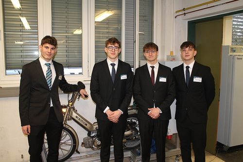 Vier junge Männer in Anzügen und Krawatten posieren neben einem Fahrrad, lächeln für die Kamera in einem Raum mit Fenstern und einer Tür.