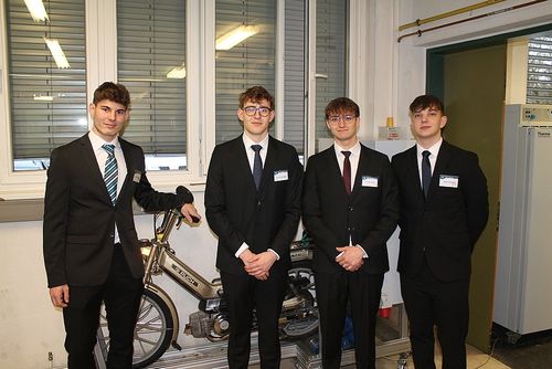 Vier junge Männer in Anzügen und Krawatten posieren neben einem Fahrrad, lächeln für die Kamera in einem Raum mit Fenstern und einer Tür.