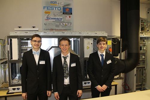 Drei Männer in Anzügen stehen vor einer Glasvitrine mit Firmenlogos. Sie lächeln und scheinen für ein Foto zu posieren.