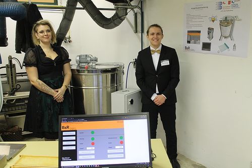 Ein Mann und eine Frau stehen neben einem großen silbernen Tank, beide lächelnd, mit einem Monitor, der einen Bildschirm vor ihnen anzeigt.
