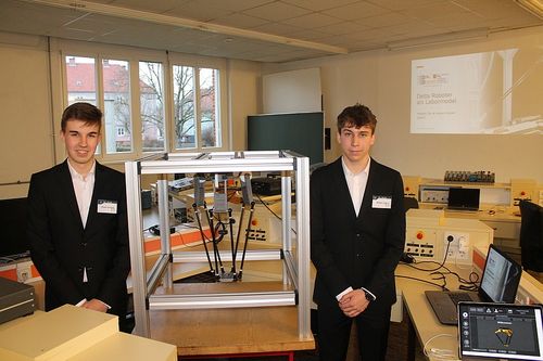Zwei Männer in Anzügen stehen in einem Labor neben einem Delta-Roboter. Laptops und andere Geräte stehen auf dem Schreibtisch.