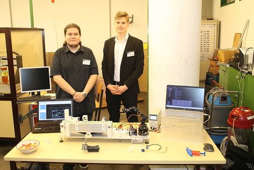 Zwei Männer stehen vor einem Tisch mit verschiedenen elektronischen Geräten, darunter Monitore und Laptops. Sie lächeln und posieren für ein Foto.