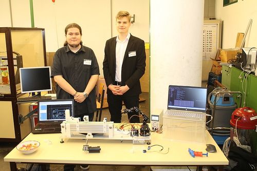 Zwei Männer stehen vor einem Tisch mit verschiedenen elektronischen Geräten, darunter Monitore und Laptops. Sie lächeln und posieren für ein Foto.