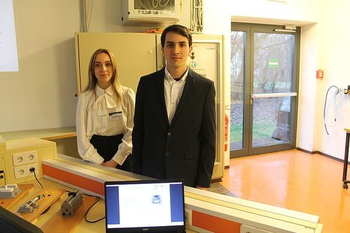 Zwei Personen stehen neben einem Laptop in einem Raum mit Glastüren, Schränken und Steckdosen.
