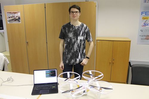 Ein Mann steht hinter einem Schreibtisch mit einem Laptop und vier Drohnen. Er trägt eine Brille und ein schwarz-weißes T-Shirt. Hinter ihm stehen Holzschränke an der Wand.