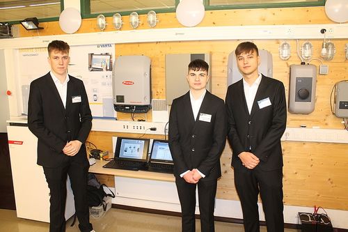 Drei Männer in Anzügen stehen vor einer Wand mit einem Stromsystem. Sie sind neben einem Schreibtisch mit zwei Laptops.