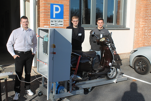 Drei Männer stehen vor einem Gebäude mit einem Roller auf einer Hebebühne, einer hält das Stromkabel. Hinter ihnen ist ein Parkplatzschild sichtbar.