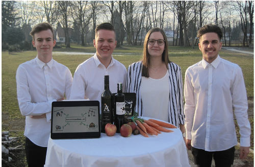 Vier Personen stehen um einen Tisch mit Obst und Gemüse. Sie lächeln und scheinen für ein Foto zu posieren. Ein Laptop steht auf dem Tisch. Dahinter befindet sich ein Rasen mit Bäumen.