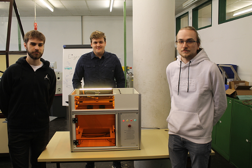 Drei Männer stehen neben einer weißen und orangefarbenen Maschine auf einem Tisch in einem Büro. Der Mann auf der linken Seite trägt eine schwarze Kapuzenjacke.