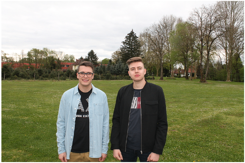 Zwei junge Männer stehen zusammen in einem Graspark. Einer trägt eine Brille und eine Jeansjacke, der andere eine schwarze Jacke. Dahinter sind Bäume und ein entferntes Gebäude.
