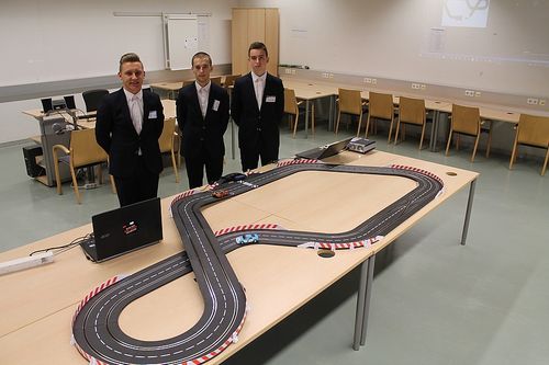 Drei Männer in Anzügen stehen um einen Tisch mit einer Spielzeugrennstrecke. Einer hält ein Auto, während die anderen zusehen. Laptops und Stühle umgeben die Szene.