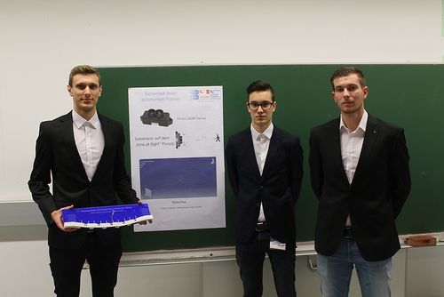 Drei Männer stehen vor einer grünen Tafel, einer hält ein blaues Modell. Die Tafel hat ein Poster mit technischen Texten und einem Diagramm.