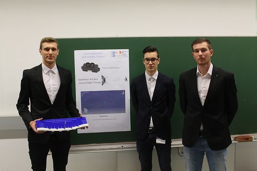 Drei Männer stehen vor einer grünen Tafel, einer hält ein blaues Modell. Die Tafel hat ein Poster mit technischen Texten und einem Diagramm.