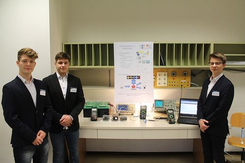 Drei Männer in Anzügen stehen vor einem weißen Tresen, mit einem großen Poster hinter ihnen. Verschiedene elektronische Geräte sind auf dem Tresen platziert, einschließlich eines Laptops.