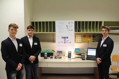 Drei Männer in Anzügen stehen vor einem weißen Tresen, mit einem großen Poster hinter ihnen. Verschiedene elektronische Geräte sind auf dem Tresen platziert, einschließlich eines Laptops.