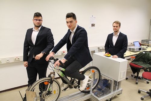 Drei Männer in Anzügen posieren mit einem Fahrrad, einer fährt es, während die anderen zuschauen, im Hintergrund ist eine weiße Box und ein roter Stuhl zu sehen.