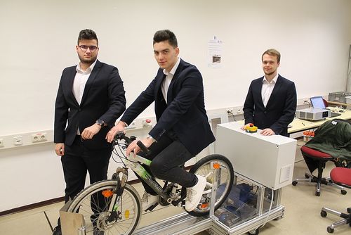 Drei Männer in Anzügen posieren mit einem Fahrrad, einer fährt es, während die anderen zuschauen, im Hintergrund ist eine weiße Box und ein roter Stuhl zu sehen.