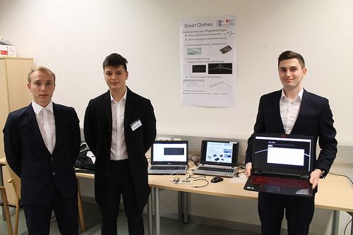 Drei Männer in Anzügen stehen neben einem Tisch mit Laptops und lächeln in die Kamera. Hinter ihnen befindet sich eine weiße Wand mit einem Poster mit dem Titel 'Smart Clothes'. Das Poster enthält Text auf Deutsch und Bilder von Laptops und Grafiken.