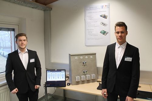 Zwei Männer in Anzügen stehen in einem Raum mit einem Schreibtisch, einem Laptop, einer Maus und einem Poster an der Wand.