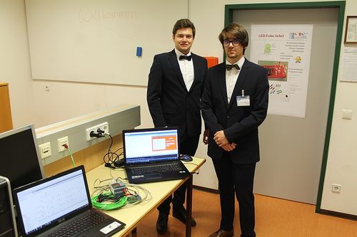 Zwei Männer in Anzügen stehen in einem Raum mit einem Schreibtisch und Laptops. Einer hat eine Maus, und eine Tafel hinter ihnen sagt Willkommen.