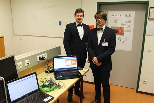 Zwei Männer in Anzügen stehen in einem Raum mit einem Schreibtisch und Laptops. Einer hat eine Maus, und eine Tafel hinter ihnen sagt Willkommen.