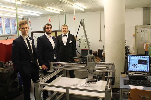 Vier Männer in Anzügen stehen in einer Werkstatt. Sie lächeln und stehen hinter einer CNC-Maschine.