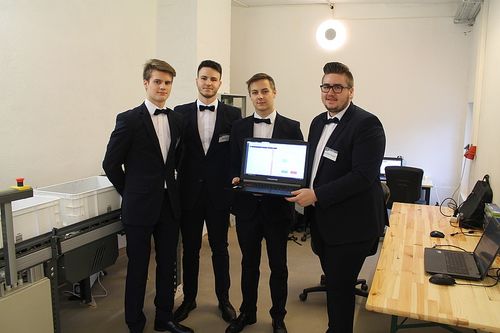 Vier junge Männer in formellen Anzügen stehen in einem Raum und einer hält einen Laptop. Sie scheinen für ein Foto zu posieren.