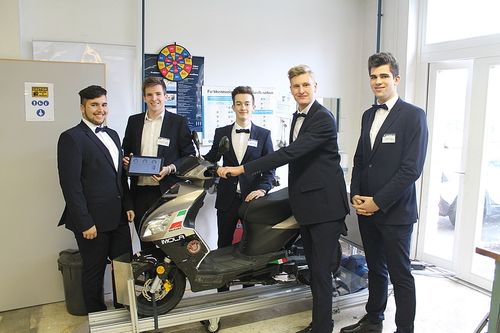 Fünf junge Männer in Anzügen und Fliegen sind um einen Roller herumstehend zu sehen und lächeln. Der Roller steht auf einer erhöhten Plattform. Hinter ihnen befindet sich eine Wand mit einer Tafel und einem Plakat.