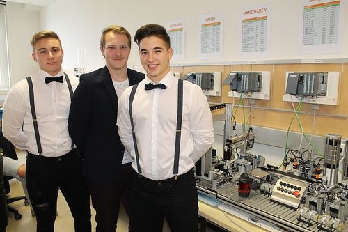Drei Männer stehen nebeneinander vor einer Maschine. Sie sind alle in weißen Hemden und Fliegen gebunden. Die Maschine hat mehrere elektronische Komponenten und Drähte, die daran angeschlossen sind. Es gibt eine weiße Wand mit einigen angehefteten Papieren.