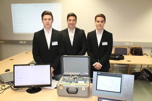 Drei Männer in Anzügen stehen in einem Büroraum mit Schreibtisch und Monitoren. Eine Aktentasche mit Kabeln und ein Bildschirm mit einem Diagramm stehen auf dem Schreibtisch.