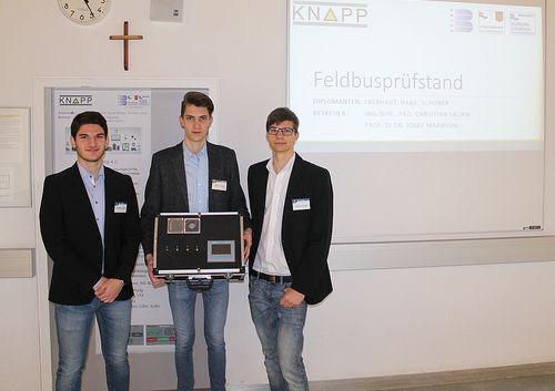 Drei Männer stehen vor einem Bildschirm und halten eine schwarze Box, möglicherweise eine Präsentation eines Projekts. Der Bildschirm zeigt 'Feldbusprufstand'. Sie tragen formelle Kleidung, einer hält eine Mappe.