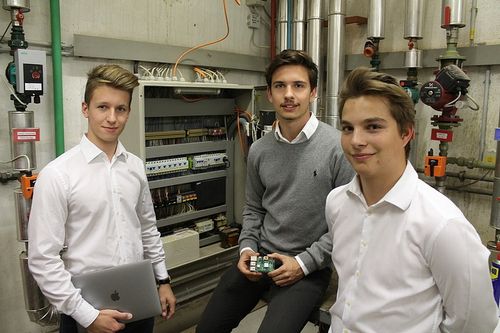 Drei junge Männer stehen zusammen in einem Raum. Der Mann auf der linken Seite hält einen Laptop, der mittlere hält ein kleines elektronisches Gerät und der rechte hat ein lächelndes Gesicht.