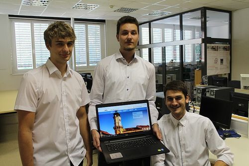 Drei junge Männer in einer modernen Büroumgebung. Der mittlere Mann hält einen Laptop mit einem Bild eines historischen Turms. Die anderen lächeln.