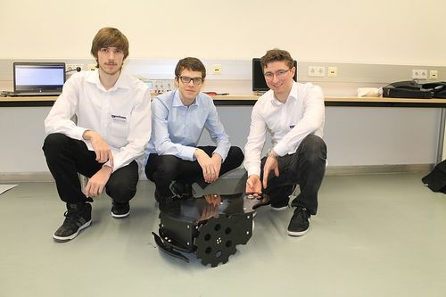 Drei Männer posieren in einer Laborumgebung, zwei kniend und einer sitzend, alle in weißen Hemden und dunkler Hose. Im Hintergrund ist ein Schreibtisch mit Monitoren und Steckdosen zu sehen.