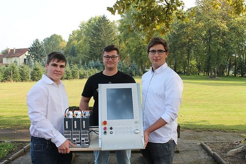 Drei Männer posieren mit einem weißen elektronischen Gerät auf einem Tisch im Freien, einer hält eine Kiste mit Ausrüstung.