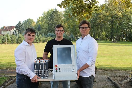 Drei Männer posieren mit einem weißen elektronischen Gerät auf einem Tisch im Freien, einer hält eine Kiste mit Ausrüstung.