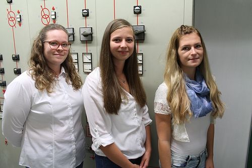 Drei Frauen stehen neben einer Wand mit elektrischen Geräten, lächelnd und posieren für ein Foto.