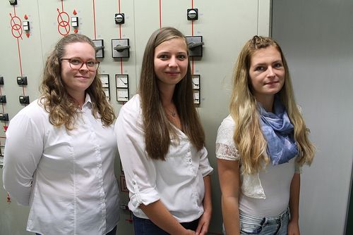 Drei Frauen stehen neben einer Wand mit elektrischen Geräten, lächelnd und posieren für ein Foto.