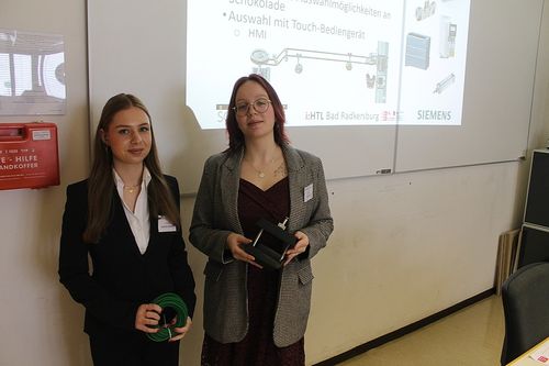 Zwei Frauen stehen vor einer Wand mit einer Projektionsleinwand. Eine hält einen grünen Schlauch, die andere ein Gerät. Siemens wird auf der Leinwand projiziert.