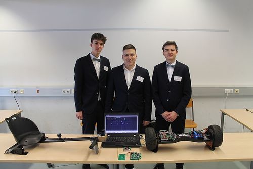 Drei Männer in Anzügen stehen in einem Klassenzimmer und lächeln. Ein Laptop steht vor ihnen auf einem Tisch, und ein kleiner Roboter ist auf dem Tisch.