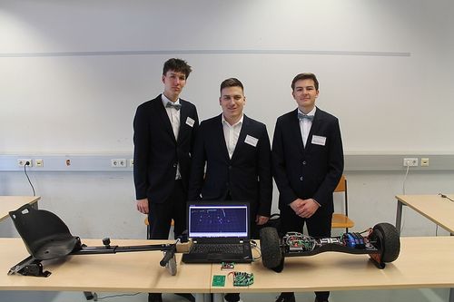Drei Männer in Anzügen stehen in einem Klassenzimmer und lächeln. Ein Laptop steht vor ihnen auf einem Tisch, und ein kleiner Roboter ist auf dem Tisch.