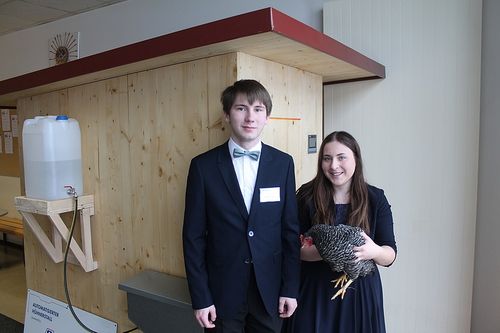 Ein junger Mann im Anzug und mit Fliege steht neben einer jungen Frau, die ein Huhn in einem Holzraum hält.