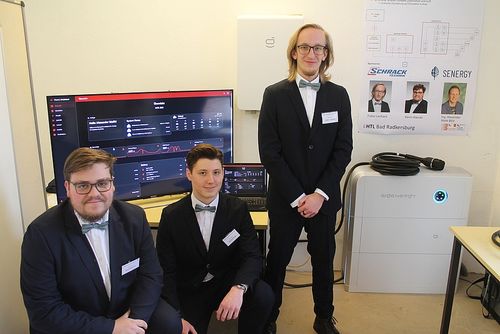 Drei Männer in Anzügen und Fliegen posieren für ein Foto mit einem großen Monitor, einem Laptop und einem weißen Netzteil.