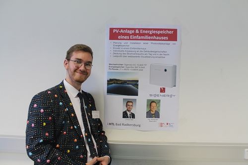 Ein Mann steht neben einem Poster mit einem Foto einer Photovoltaikanlage. Das Poster hat Text auf Deutsch, ein Foto von zwei Männern und ein Foto einer Solaranlage.