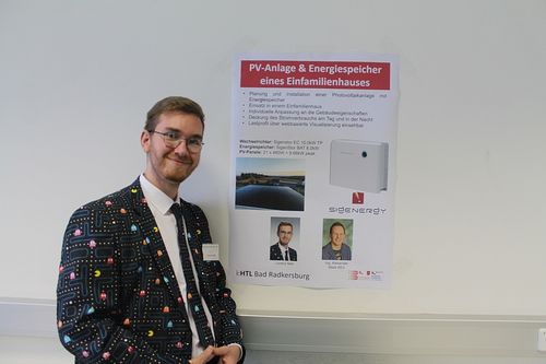Ein Mann steht neben einem Poster mit einem Foto einer Photovoltaikanlage. Das Poster hat Text auf Deutsch, ein Foto von zwei Männern und ein Foto einer Solaranlage.